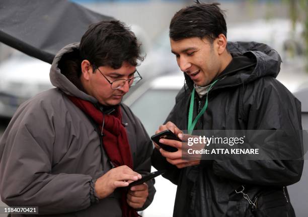 Bike-delivery food messenger stands next to another man as they both use smartphones along the side of a street in the Iranian capital Tehran on...