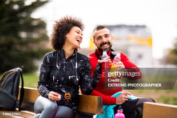 zwei junge sportler in sportbekleidung setzen ihre energy-drink-flaschen zusammen, während sie auf einer bank in einem öffentlichen park sitzen - energydrink stock-fotos und bilder
