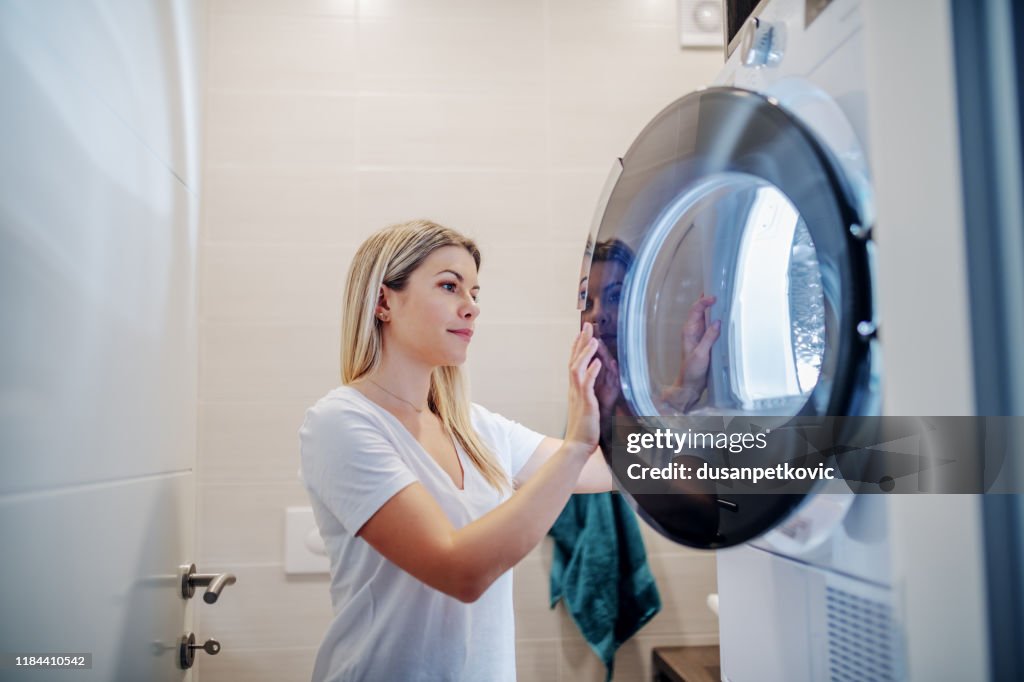 Worthy caucasian young blond housewife closing drying machine while standing in bathroom in late hours.