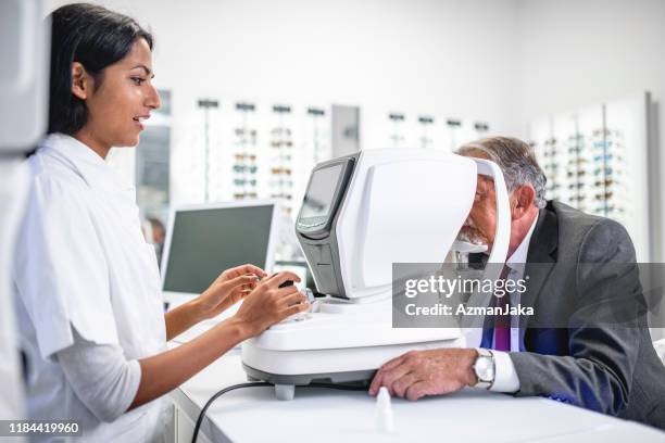 senior man getting an eye exam and talking to the indian female doctor in her clinic - ophthalmologist stock pictures, royalty-free photos & images
