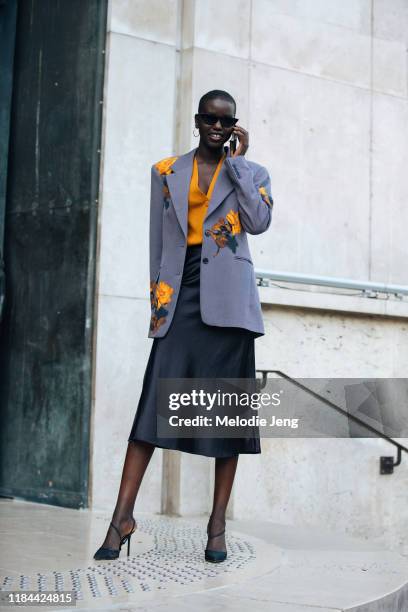 Model Adut Akech talks on the phone and wears a purple Dries Van Noten blazer with floral embellishments, an orange button-up cardigan top, a black...