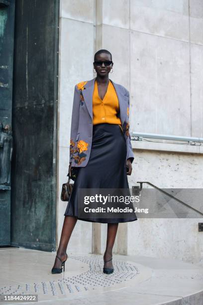 Model Adut Akech wears a purple Dries Van Noten blazer with floral embellishments, an orange button-up cardigan top, a black knee-length silk skirt,...