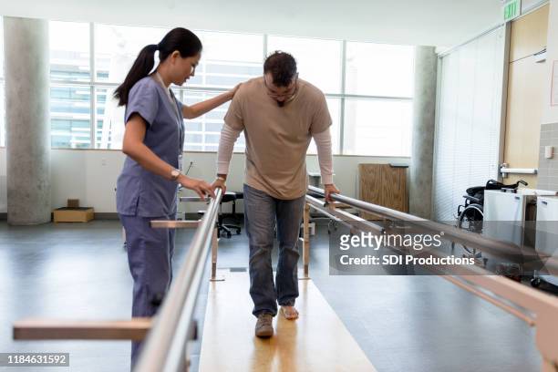 el paciente masculino toma los primeros pasos usando barras paralelas ortopédicas - fisioterapia fotografías e imágenes de stock