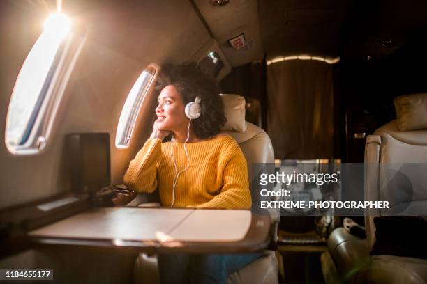 joven mujer moderna sentada en un jet privado, escuchando música a través de los auriculares y mirando a través de la ventana - first class fotografías e imágenes de stock