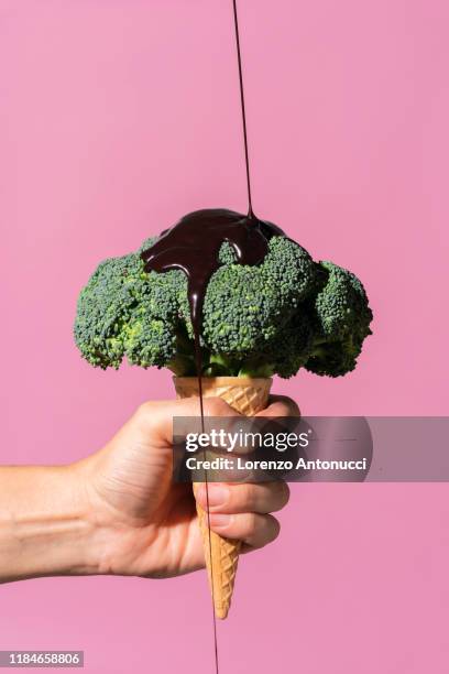 studio shot of man's hand holding ice cream cone with broccoli on top and pouring chocolate sauce, against pink background - broccoli stock pictures, royalty-free photos & images