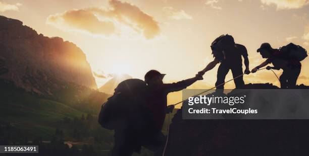 young asian three hikers climbing up on the peak of mountain near mountain. people helping each other hike up a mountain at sunrise. giving a helping hand. climbing. helps and team work concept - herausforderung stock-fotos und bilder