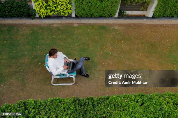 a high angle view of a young adult handsome boy using laptop in the lawn while sitting on an outdoor chair - man-sitting-on-grass-using-laptop-elevated-view stock pictures, royalty-free photos & images
