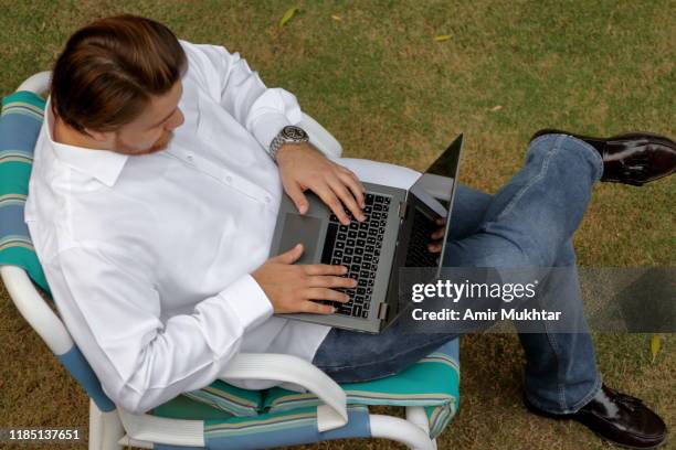 a high angle view of a young adult handsome boy using laptop in the lawn while sitting on an outdoor chair - man-sitting-on-grass-using-laptop-elevated-view stock pictures, royalty-free photos & images