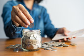 The woman hand is putting a coin in a glass  bottle and a pile of coins on a brown wooden table,Investment business, retirement, finance and saving money for future concept.