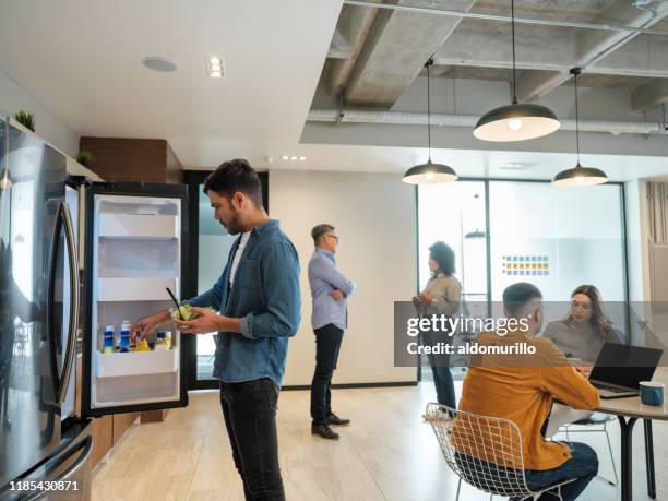 young latin man standing in front of fridge in lunch room - office lunch stock pictures, royalty-free photos & images