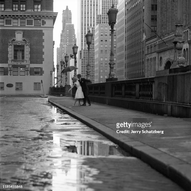 In the early morning, a female model, in a white dress, embraces a male model, in a suit, under an umbrella on sidewalk of the Grand Central Station...