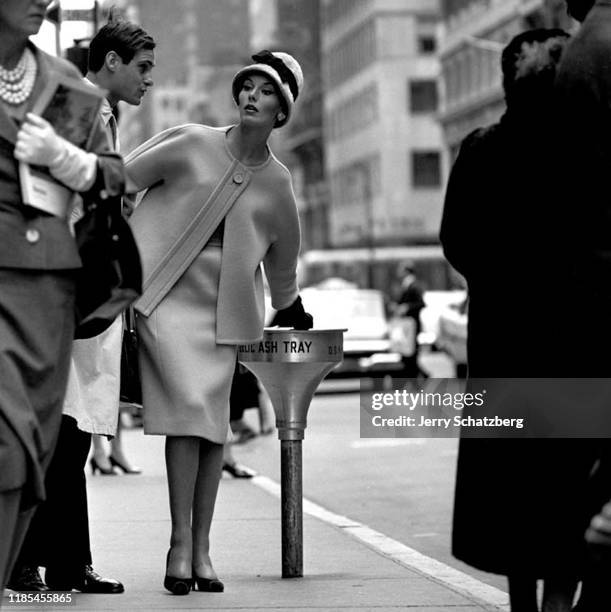 In a white hat with a black bow and a skirt suit, American fashion model Carmen Dell'Orefice puts out a cigarette in a public ash tray on a crowded...