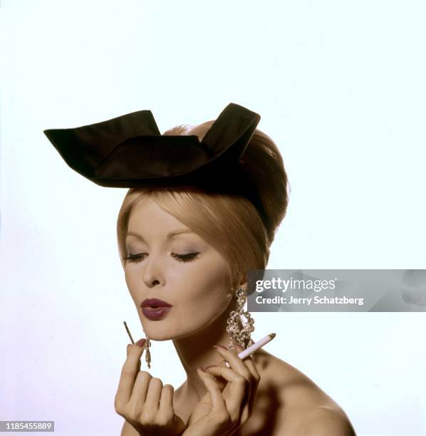 View of fashion model Sara Thom, in a black, bow hat and jeweled earrings, as she blows out a match in one hand and holds a lit cigarette in the...