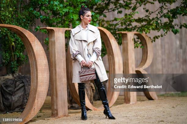 Isis Valverde wears earrings, a beige Dior trench coat, a Dior book tote bag, shiny patent leather pointy kitten-heeled thigh-high boots, outside...