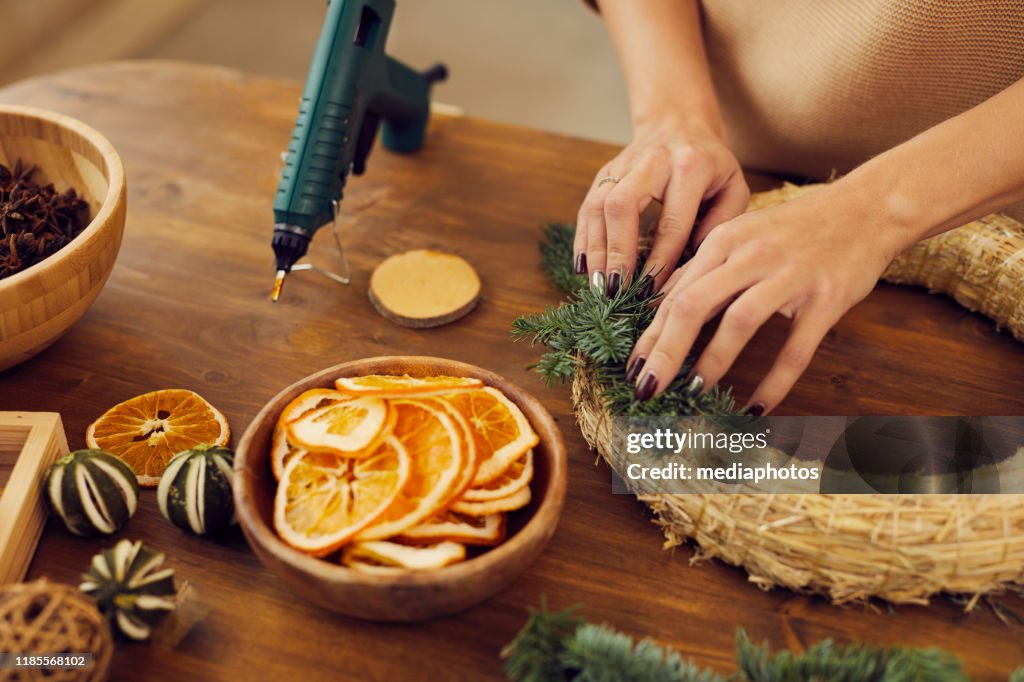 Close-up of unrecognizable woman with manicure standing at wooden table in workshop and using hot glue while attaching fir tree twigs to wreath base