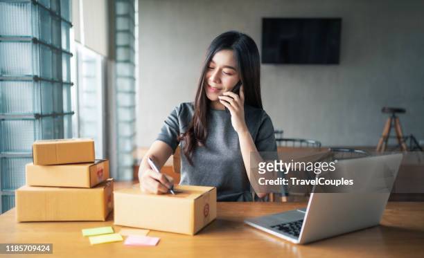 young business woman working selling online. entrepreneur owner using smartphone or laptop taking receive and checking online purchase shopping order to preparing pack product box. packing goods for delivery to customer. e-commerce. online shopping - e commerce 個照片及圖片檔