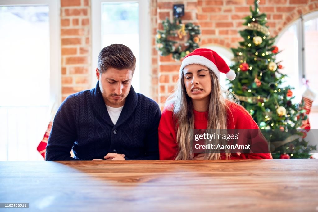 Young couple wearing santa claus hat sitting on chair and table around christmas tree at home with hand on stomach because nausea, painful disease feeling unwell. Ache concept.