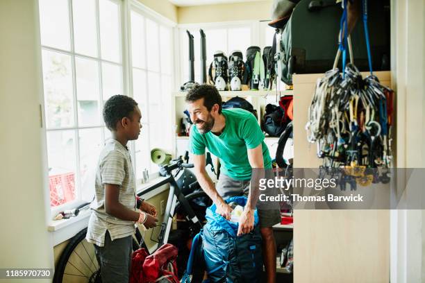 smiling father packing backpack with son in gear room in home - packing kids backpack stockfoto's en -beelden