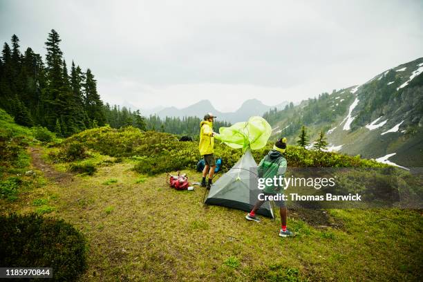 father putting rainfly on tent during backpacking trip in rain with son - tough love stock-fotos und bilder