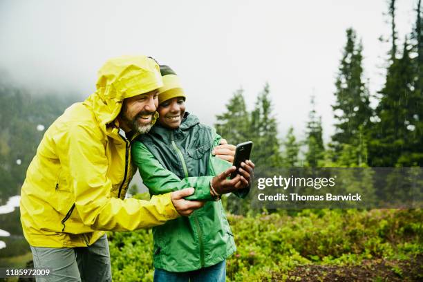 smiling father and son taking selfie with smart phone while camping during rainstorm - grüne jacke stock-fotos und bilder