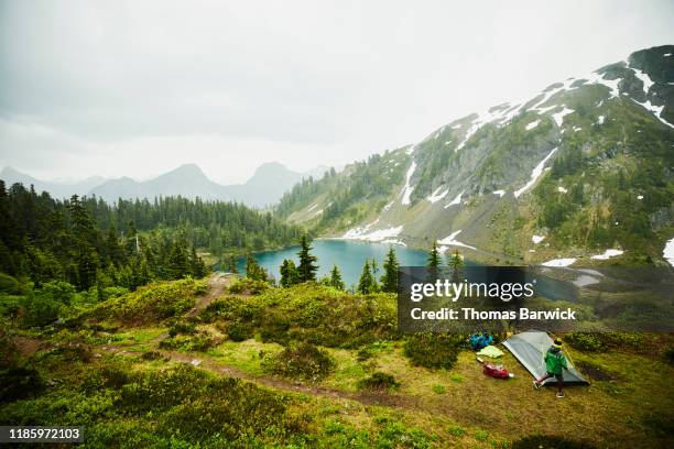 father and son setting up tent above alpine lake during backpacking trip in rain - american influenced fotografías e imágenes de stock