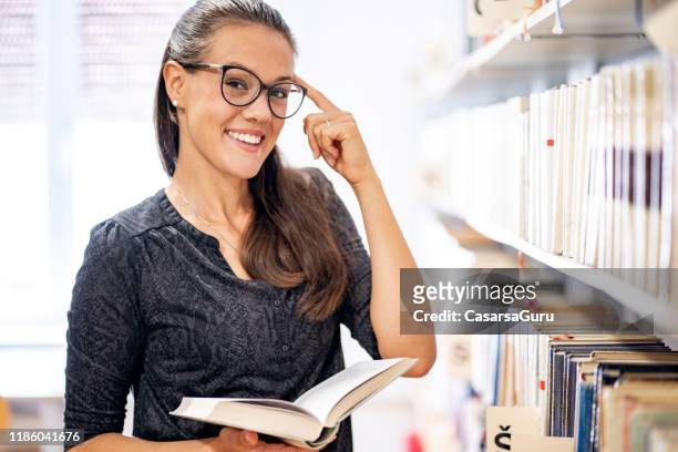 portrait of smiling young woman standing in library and pointing at her head holding a book - muster point stock pictures, royalty-free photos & images
