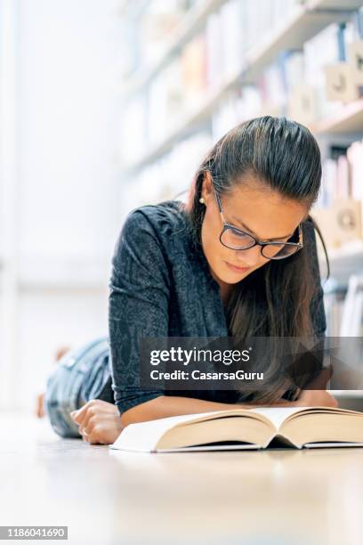 beautiful young woman lying on floor in library and reading a book - thick reading glasses stock pictures, royalty-free photos & images