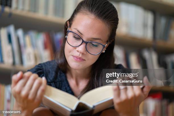 young woman reading a thick book in library with her reading glasses - thick reading glasses stock pictures, royalty-free photos & images