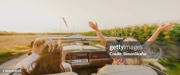 two women with raised arms driving convertible car on country road - convertible stock pictures, royalty-free photos & images