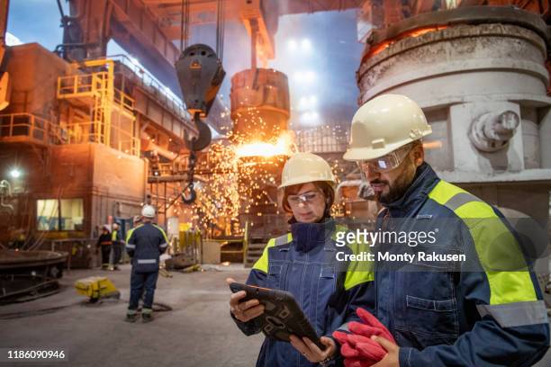 male and female steelworkers using digital tablet during steel pour in steelworks - stahlindustrie stock-fotos und bilder