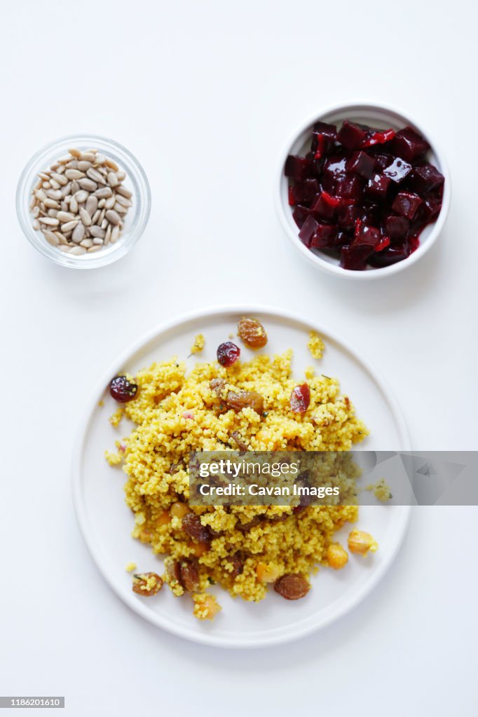 Overhead view of couscous served in plate with beetroot salad and seeds in bowls on white background