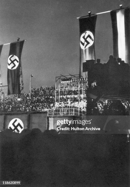 Flags bearing the swastika logo of the Nazi Party hang at a rally with crowds of people in attendance, with party leader Adolf Hitler standing on a...