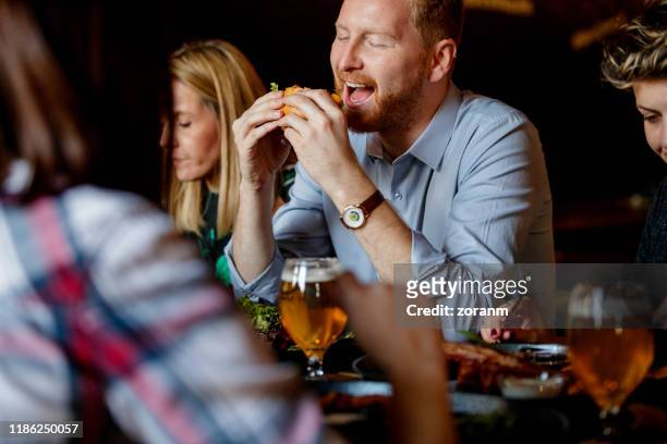 young redhead man with friends in a pub eating hamburger with pleasure - bbq sandwich stock pictures, royalty-free photos & images