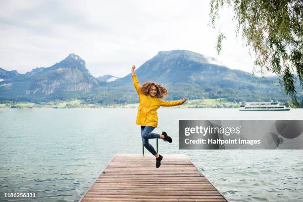 front view of young woman tourist standing by lake on a hiking trip, jumping. - frau mit gelben regenmantel stock-fotos und bilder