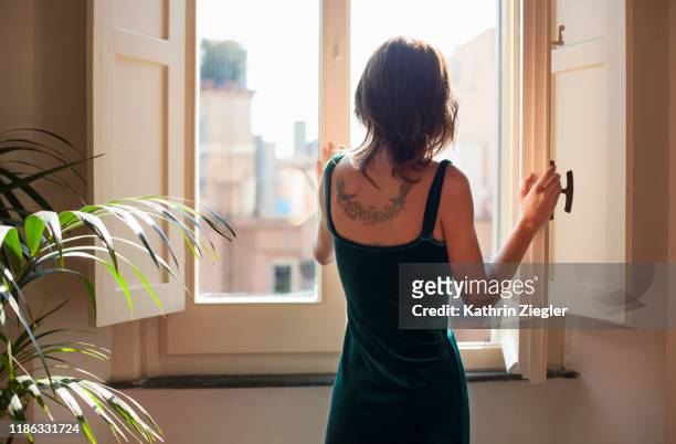 young woman in velvet dress opening window, rear view showing tattoo - lüften stock-fotos und bilder