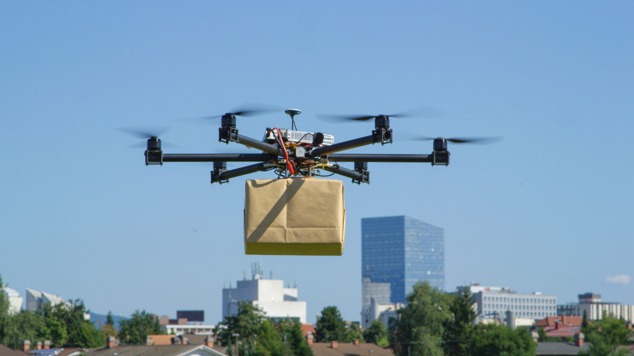CLOSE UP: UAV drone delivery delivering big brown post package into urban city CLOSE UP: UAV drone delivery delivering big brown post package into urban city
