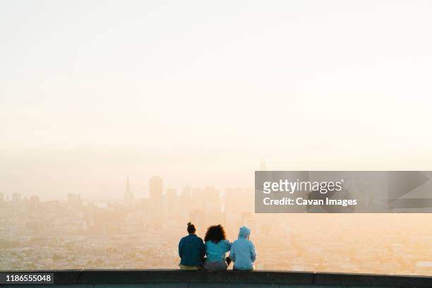 rear view of female friends looking at cityscape while sitting against clear sky during sunset - back shot position stock pictures, royalty-free photos & images