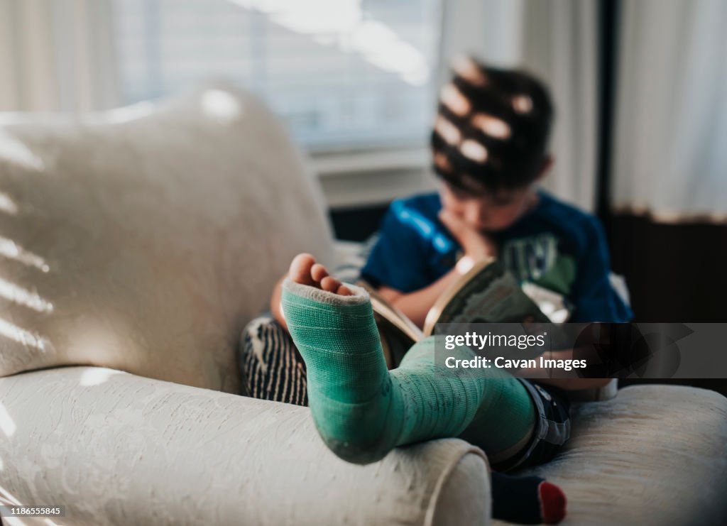 Boy with broken leg reading book while relaxing on armchair at home