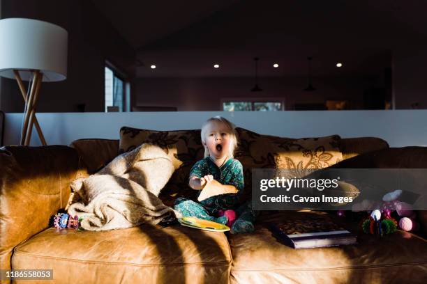 cute girl yawning while eating bread on sofa at home - yawning stock photos et images de collection