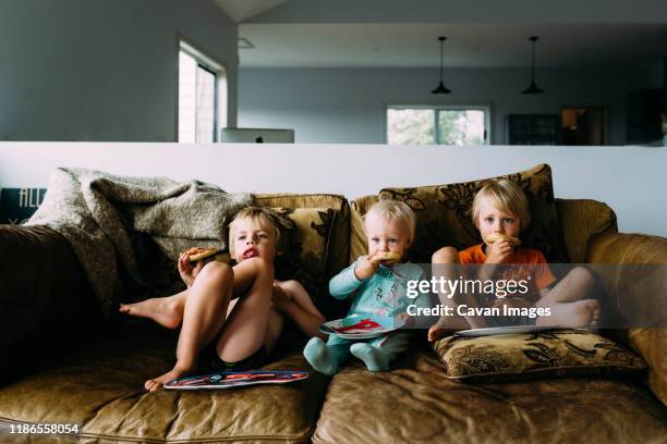 siblings eating snacks watching tv while relaxing on sofa at home - australian family home stockfoto's en -beelden