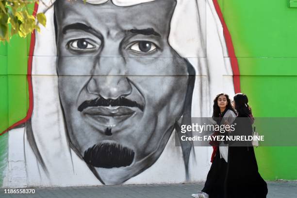 Saudi women walk past a mural painting showing King Salman bin Abdulaziz on Tahliya street in the capital Riyadh on December 5, 2019.