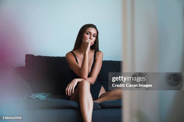 portrait of seductive young woman wearing black one piece swimsuit while sitting on sofa at home - 魅惑的な女性 ストックフォトと画像