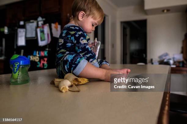 young boy sitting on kitchen counter with banana, cup, and pouch - pouch stock pictures, royalty-free photos & images