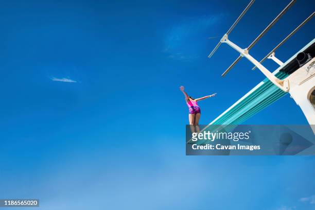 low angle view of carefree girl standing on diving platform against blue sky during sunny day - jumping girl stock-fotos und bilder