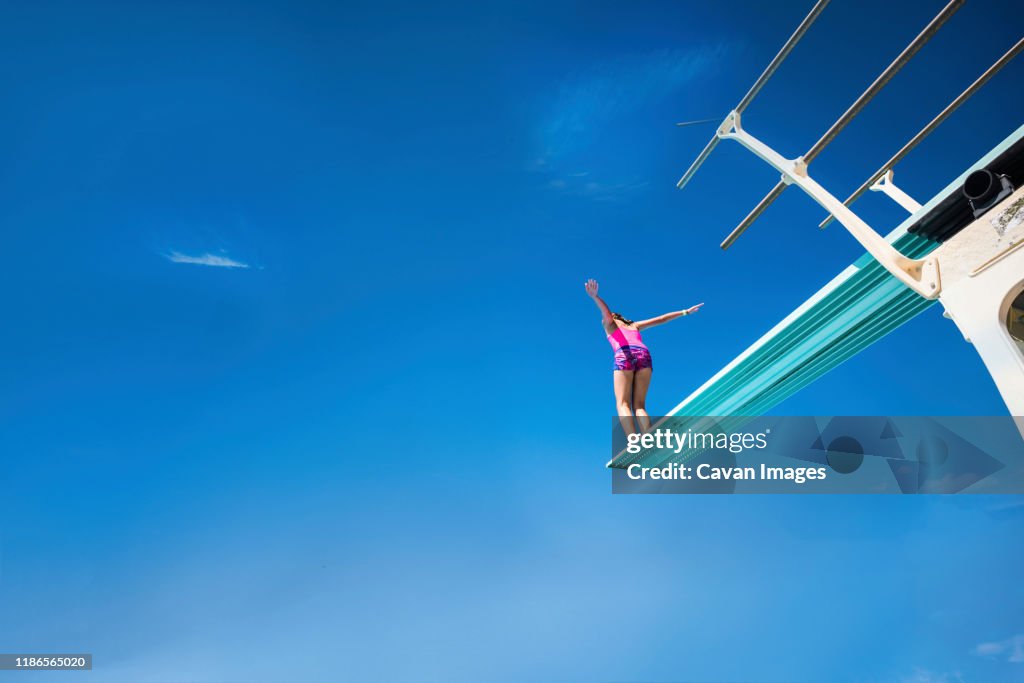Low angle view of carefree girl standing on diving platform against blue sky during sunny day