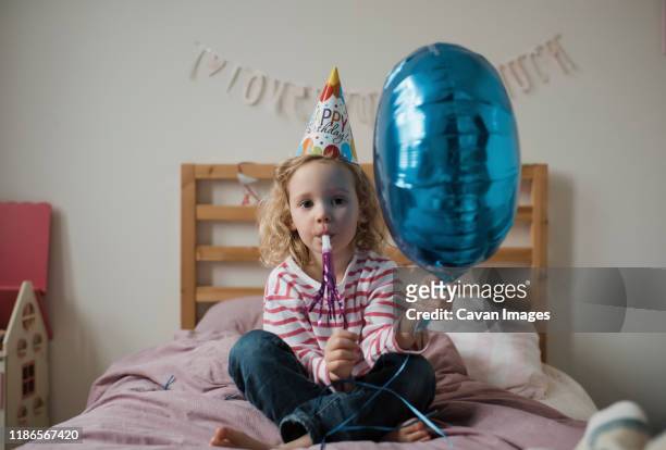 portrait of cute girl playing with party horn blower and balloon while sitting on bed against wall at home - party horn blower imagens e fotografias de stock