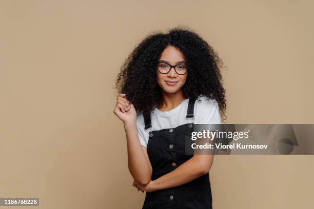Isolated shot of pleasant looking young African American woman touches curly hair, has bushy hairstyle, wears optical glasses, white t shirt and black overalls, poses over brown studio background
