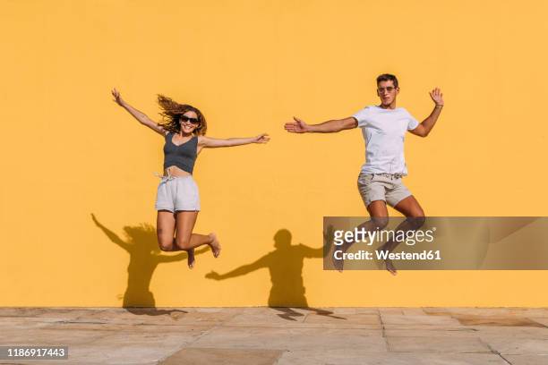 young couple jumping in front of a yellow wall - barefoot couples stock pictures, royalty-free photos & images