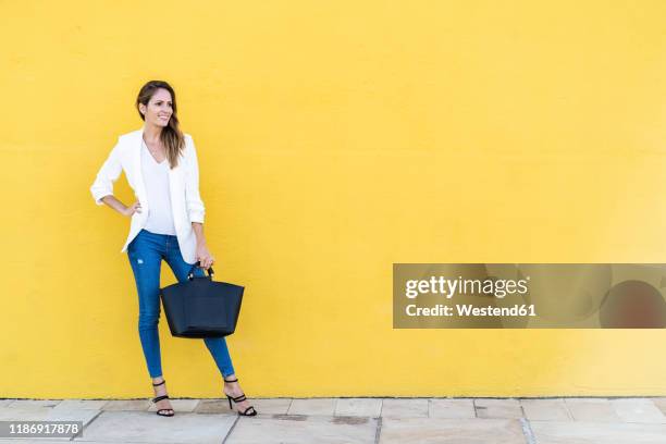 smiling woman standing at a yellow wall holding a handbag - bolso marrón fotografías e imágenes de stock