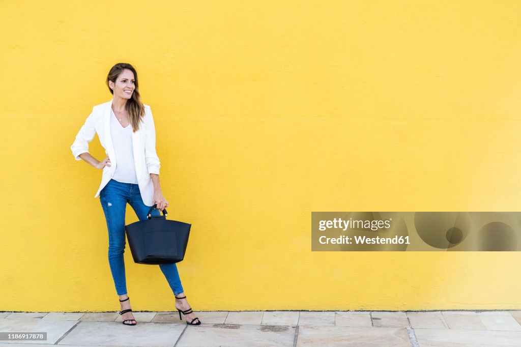 Smiling woman standing at a yellow wall holding a handbag